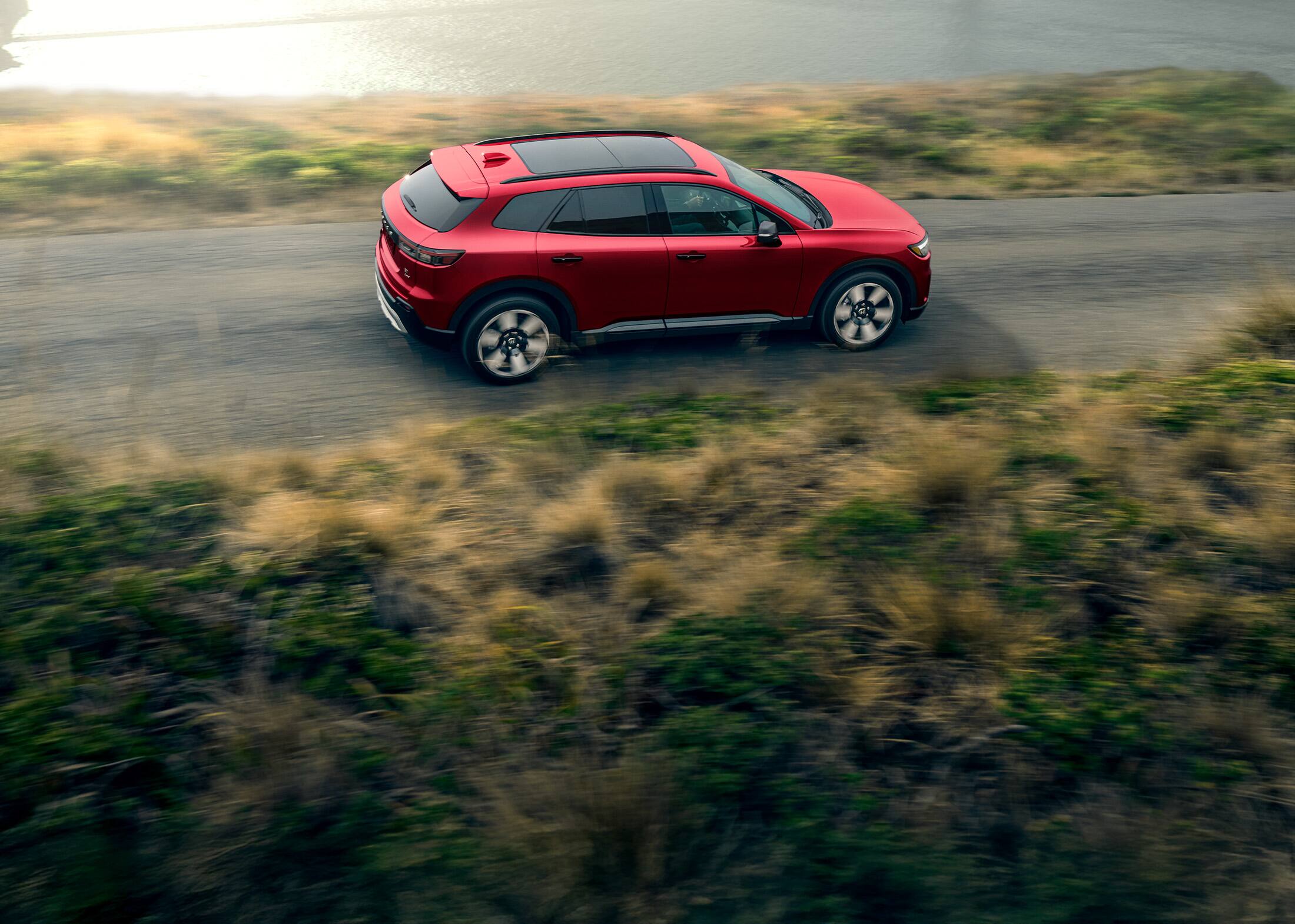 Rear passenger view of the 2024 Honda Prologue Elite AWD in Scarlet Red Metallic, shown driving with a large bridge in the background.