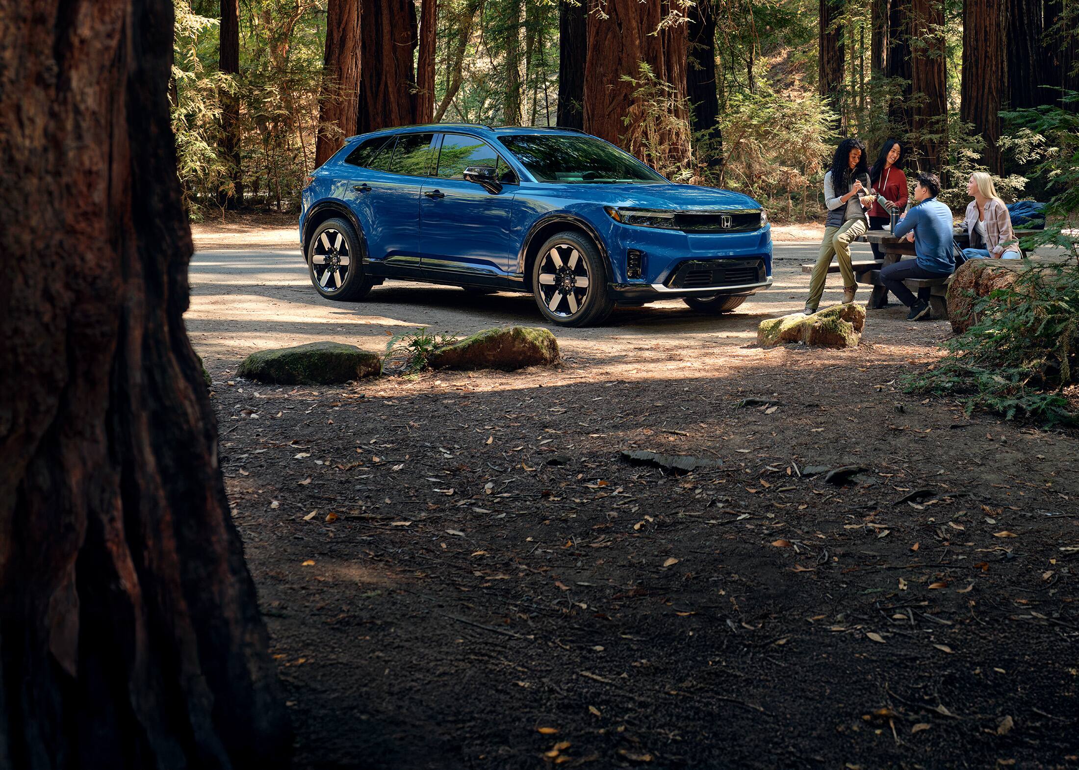 Front passenger view of the 2024 Honda Prologue Elite in Pacific Blue Metallic, shown parked with redwood trees behind it